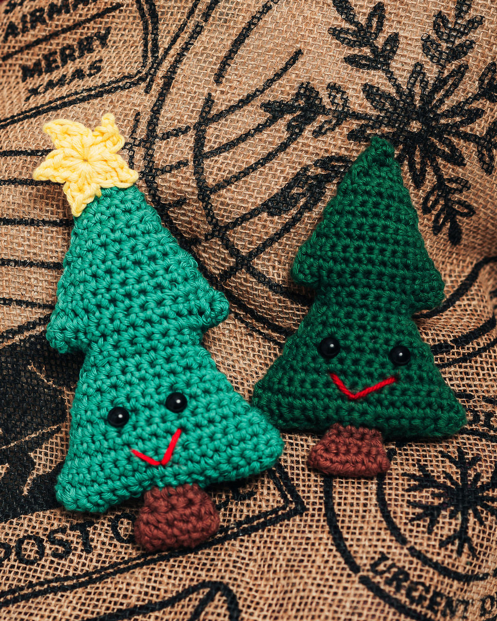 Two green crocheted Christmas trees with facial expressions on a textured brown background.